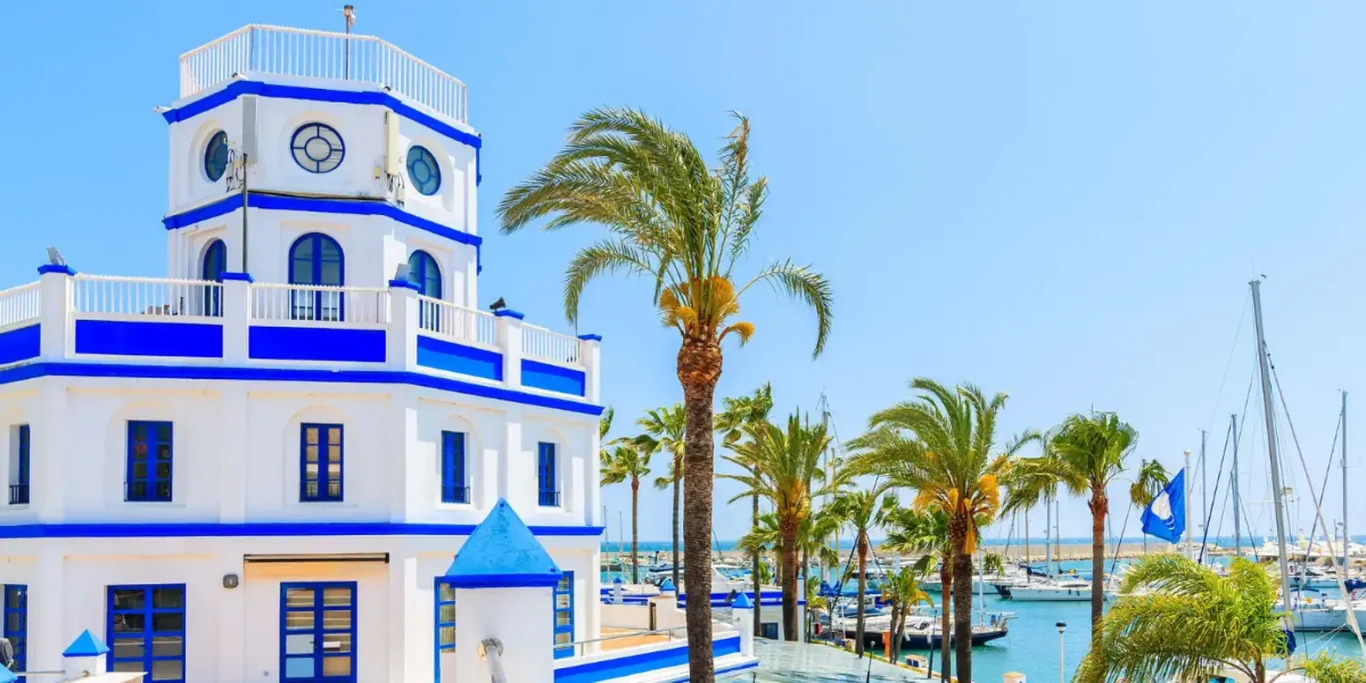 Edificio blanco y azul junto a palmeras y un puerto lleno de yates.