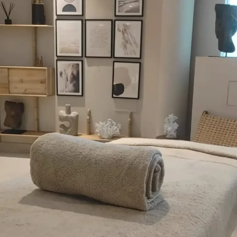 Towel rolled up on a massage table, with a modern lounge and plants in the background. The ideal setting to enjoy a massage at home in your 