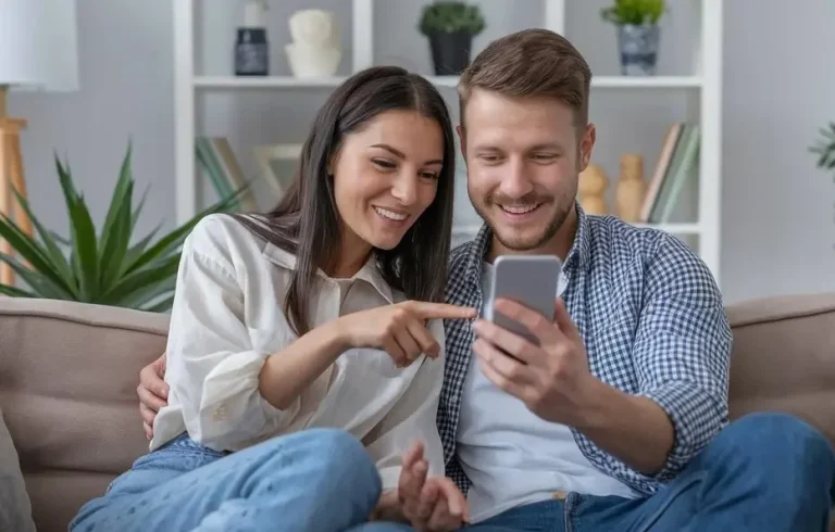 Young couple sitting on the sofa, laughing and looking at the phone, choosing their home massage in Marbella.