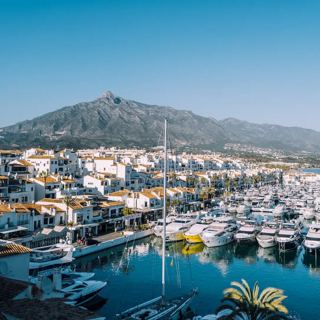 Harbour with boats, white buildings and mountains under a blue sky with clouds.