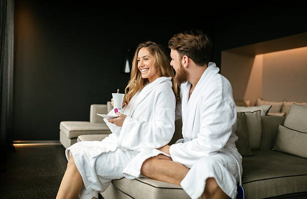 Smiling couple in white robes, sitting on a sofa with drinks in their hands after receiving a massage at home in Marbella
