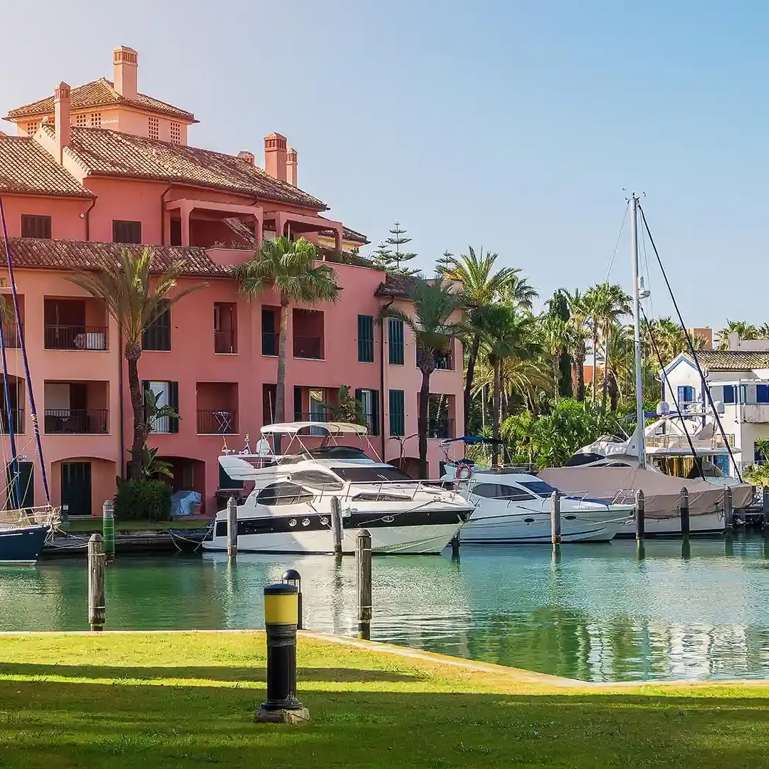 Colourful canal-side buildings with yachts and palm trees in a sunny setting.