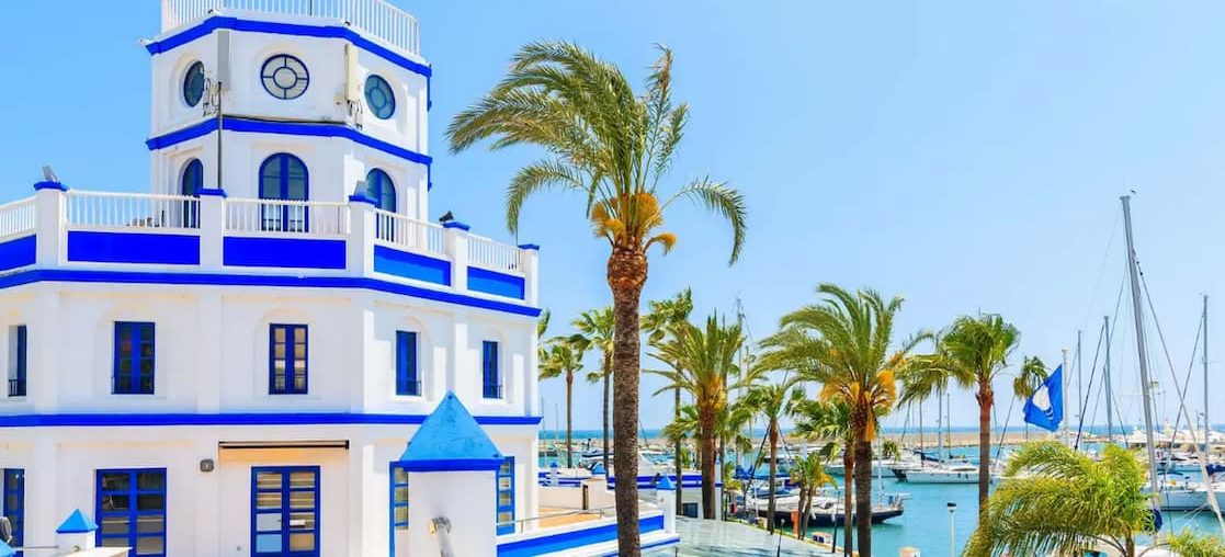 Movil massage in Estepona White and blue building next to Estepona marina with palm trees and boats in the background.