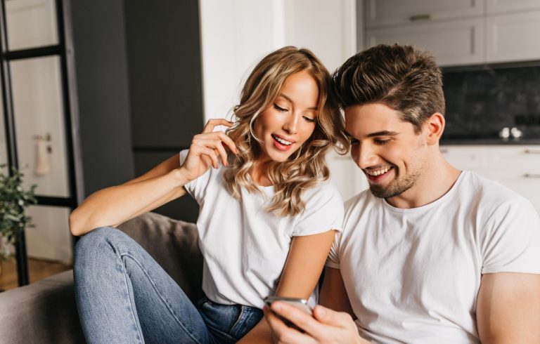 Young couple sitting on the sofa, laughing and looking at the phone, choosing their home massage in Marbella.