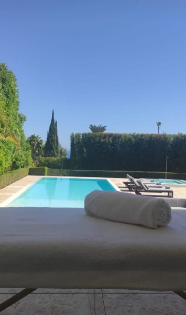View of a pool surrounded by vegetation and sun loungers under a clear blue sky.