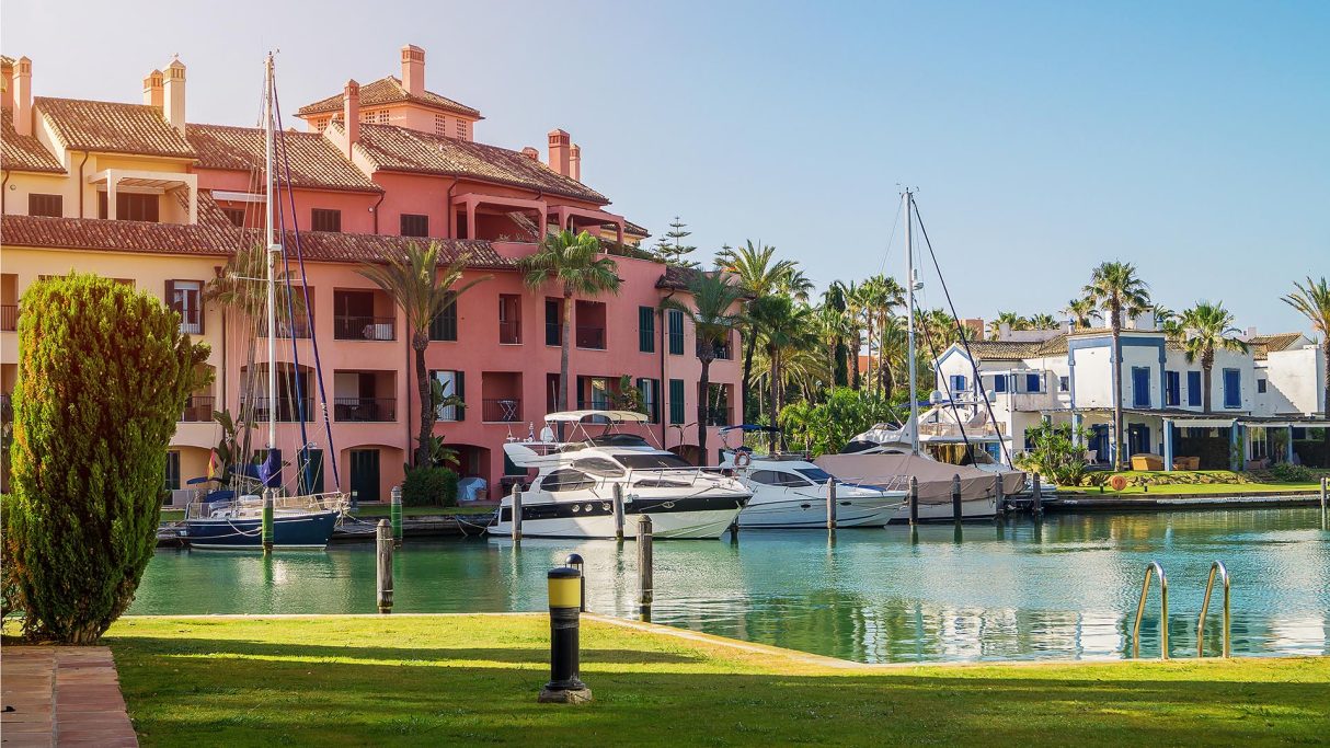 Colourful buildings next to a port with yachts and palm trees on a sunny day.Enjoy the experience of a massage at home in Sotogrande Spain.