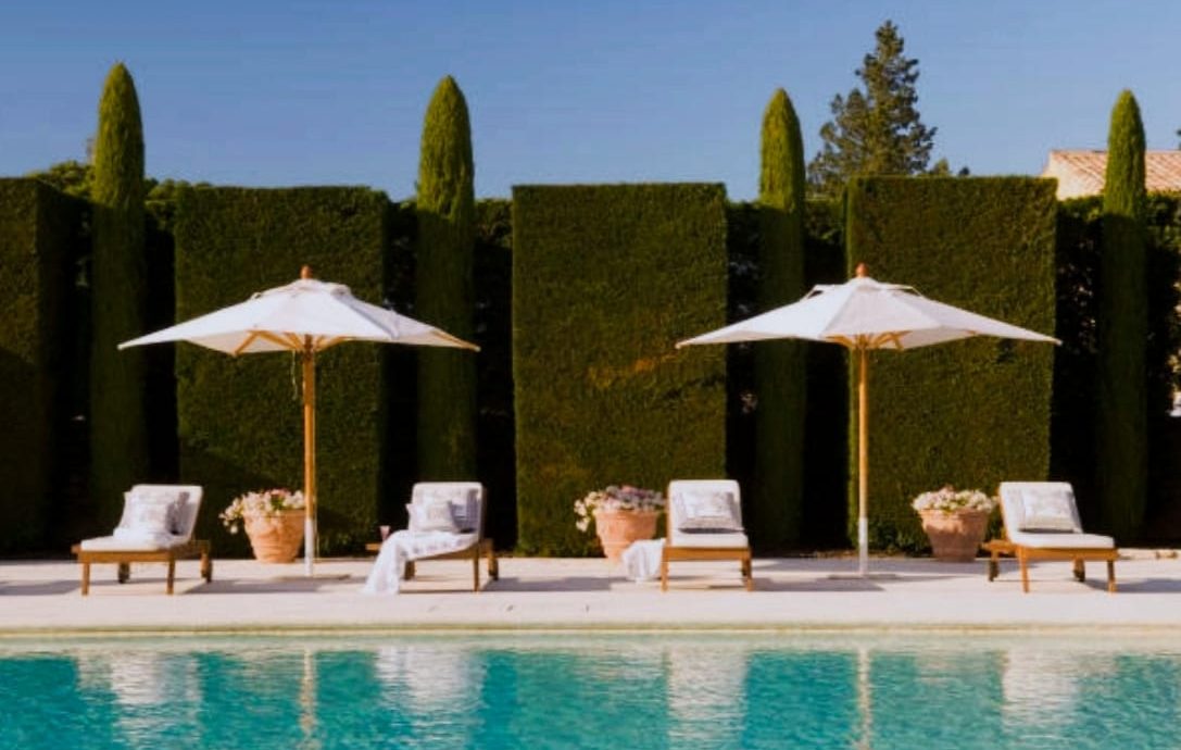 Sun umbrellas and lounge chairs next to a swimming pool surrounded by tall green hedges.