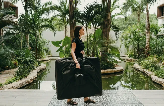 Person carrying a large briefcase in a tropical setting with palm trees and water.